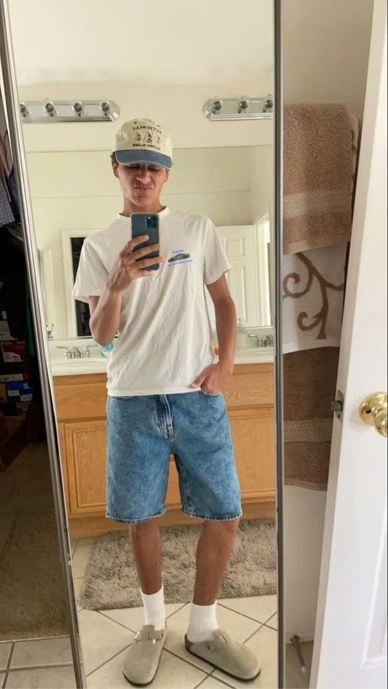 Person taking a mirror selfie wearing a white t-shirt, blue shorts, and a cap.